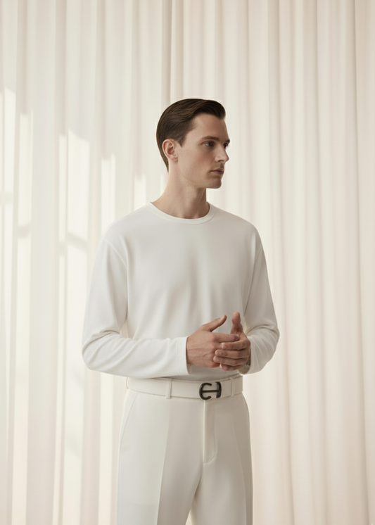 Man wearing a white long-sleeve shirt and pants with a belt against a light curtain background